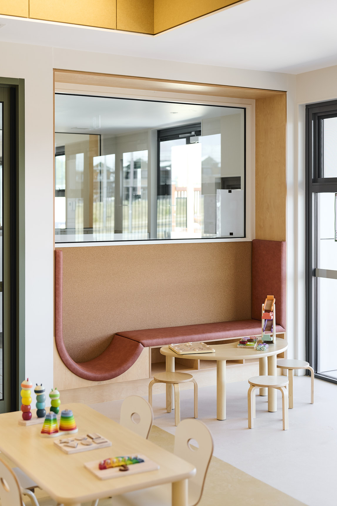 Contemporary early‑learning classroom featuring child‑sized wooden tables and chairs, colourful educational toys, puzzles, and stacking rings. Includes a built‑in seating nook and large windows creating a bright, welcoming preschool environment.