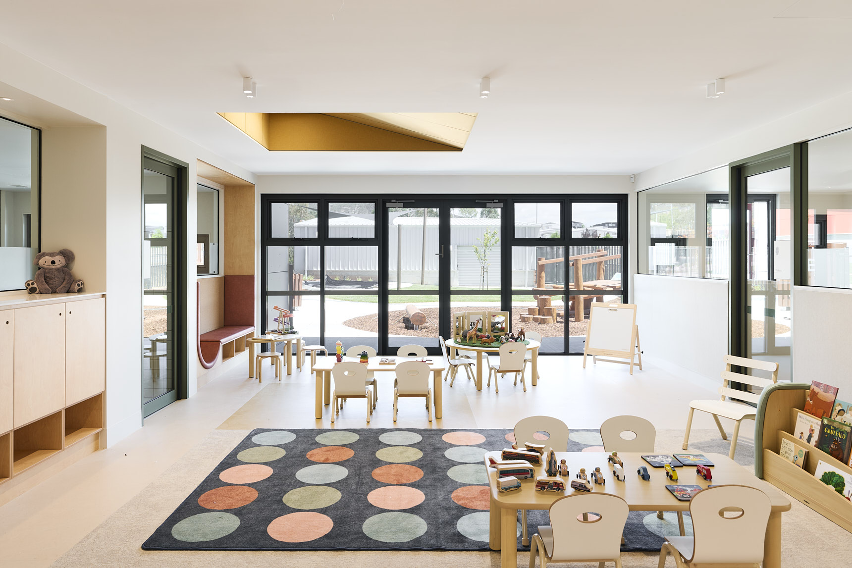Light‑filled early childhood classroom with small tables, art supplies, books, toys, and a colourful circular rug. Large windows open to an outdoor play area with wooden climbing structures, creating an engaging learning space for young children.