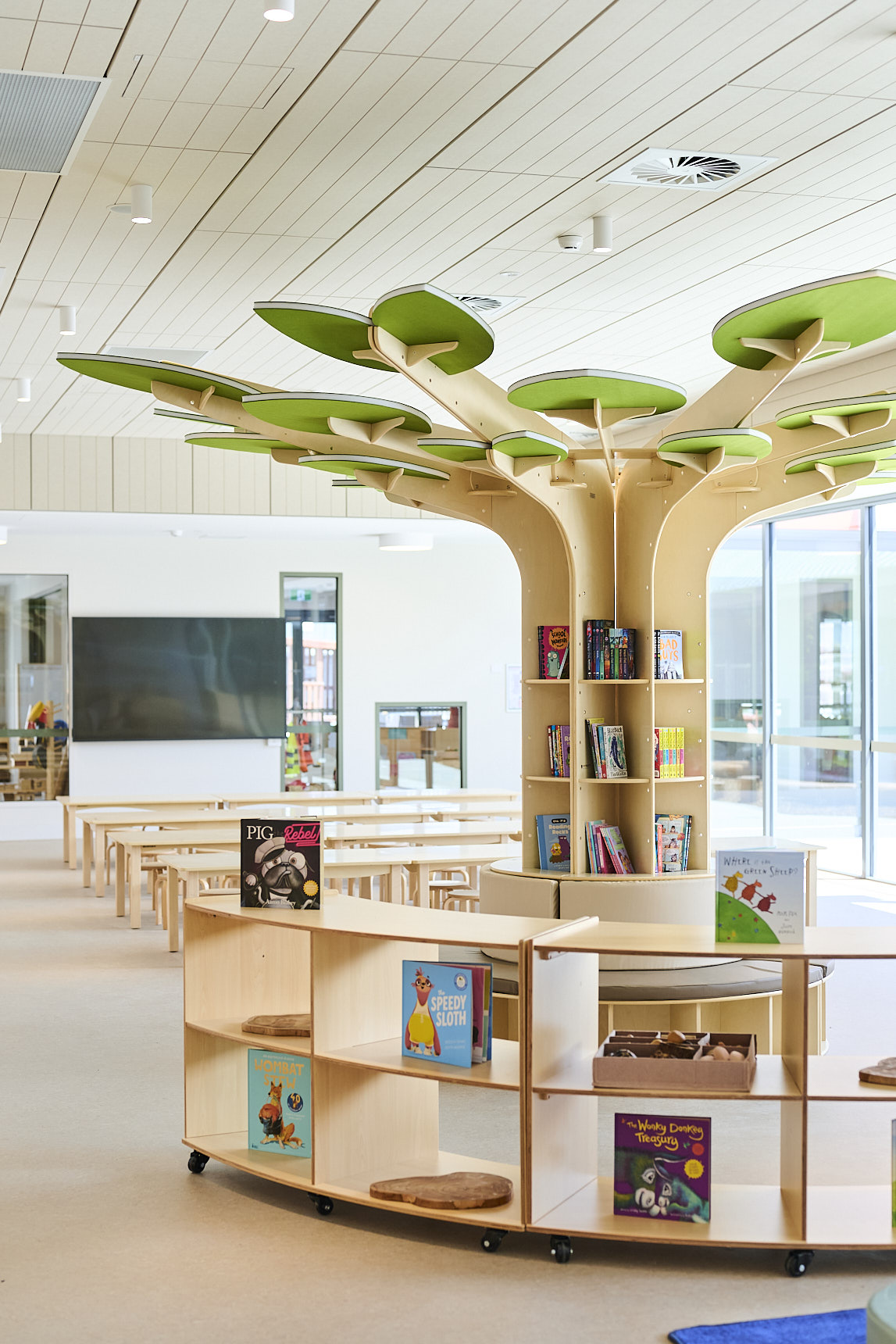 Modern children’s library with a large tree‑shaped bookshelf, curved wooden book displays, colourful picture books, and bright natural light. Features tables, stools, and an inviting reading area designed for early‑learning environments.