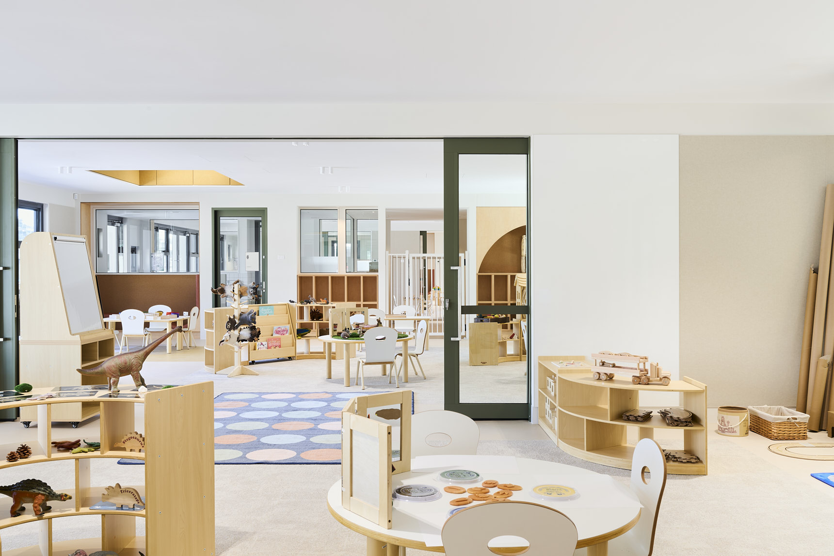 Open preschool classroom with round wooden tables, child‑sized chairs, natural play materials, wooden blocks, toy dinosaurs, and a colourful circular rug. Glass doors reveal an adjoining learning space, promoting exploration and play‑based learning.